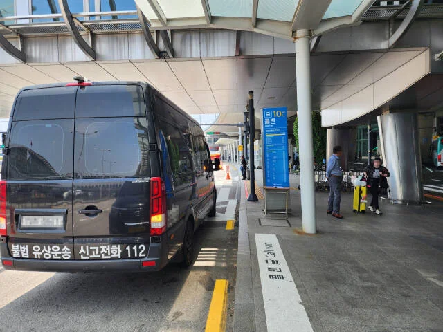 空港の前に止まる黒いワゴン車。後ろに「불법 유상운송 신고전화 112」と書かれている
