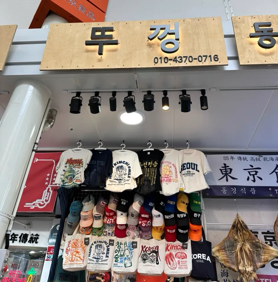 木の看板に「뚜껑」と書かれたANYWEARの店先。帽子やTシャツ、トートバッグが掛かっている