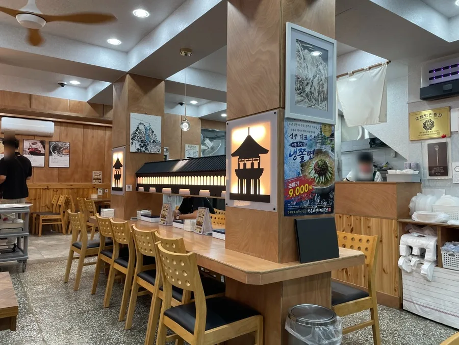 パク・ヨンジャ慶州明洞チョルミョン 本店 — 木目のカウンターとテーブル席